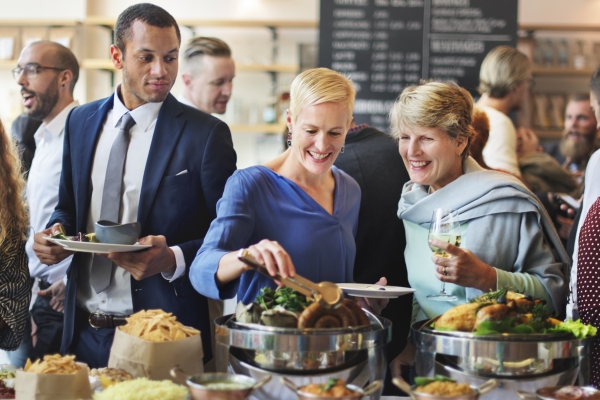 A dozen smartly dressed people mingling at an indoors event. In the foreground are four white woman and one mixed race man holding plates and glasses and reaching into a full buffet of assorted food